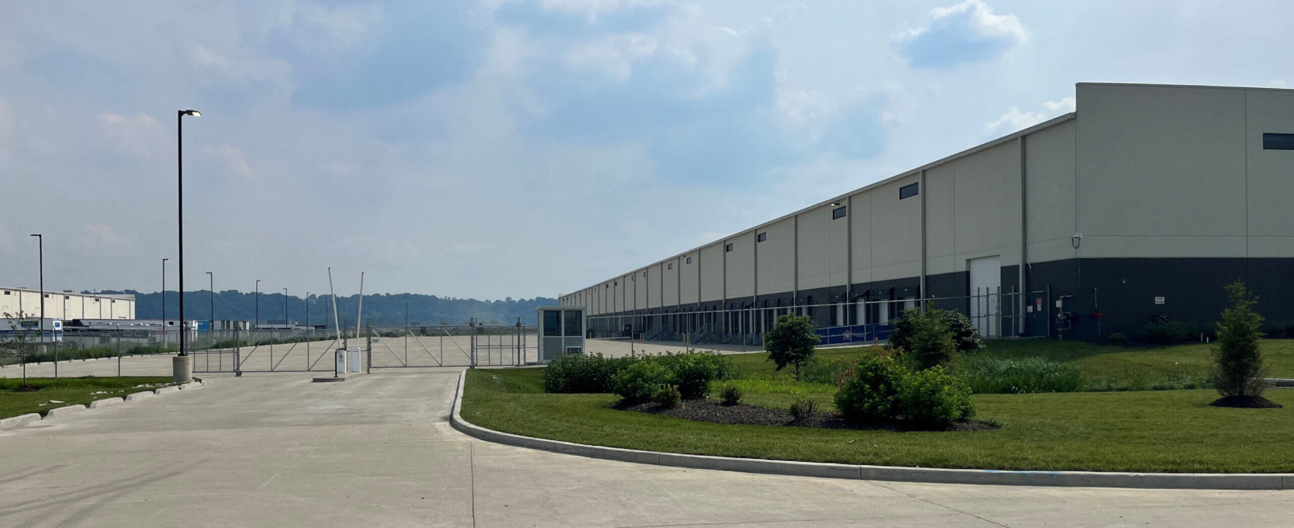New CLS Location Docks A wide concrete driveway leads to a fenced gate beside a large, beige industrial warehouse on a cloudy day, with small shrubs and grass bordering the road.